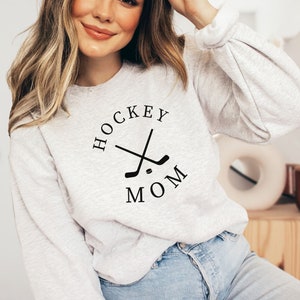May include: A light gray sweatshirt with the text "HOCKEY MOM" in a circular design with crossed hockey sticks. The sweatshirt has long sleeves and is made of a soft material.