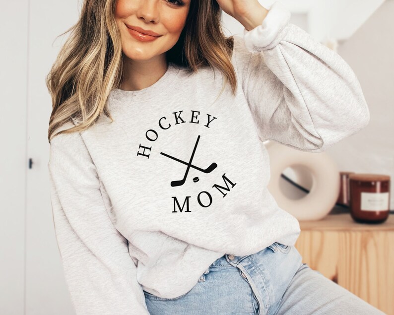 May include: A light gray sweatshirt with the text "HOCKEY MOM" in a circular design with crossed hockey sticks. The sweatshirt has long sleeves and is made of a soft material.