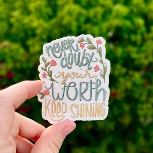May include: A white sticker with the words "Never doubt your WORTH Keep SHINING" in a script font. The words are in teal and gold, with floral accents in pink and green. The sticker is held in a hand against a blurred green background.