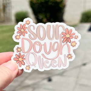 May include: A white sticker with pink and peach lettering that reads "Sour Dough Queen". The sticker is decorated with pink and peach flowers.