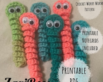 May include: Five crocheted worry worm toys in green, teal, and pink with googly eyes. The worms are arranged in a semi-circle. The text "CROCHET WORRY WORM PATTERN", "PRINTABLE NOTECARDS INCLUDED", "ZariBees", and "PRINTABLE PDF" is visible in the image.