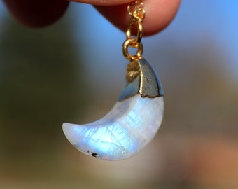 May include: A crescent moon pendant necklace. The moon-shaped pendant is made of iridescent, opalescent stone with a gold-colored metal cap and chain. The pendant is held up against a blurred background.