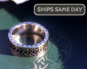 May include: A silver ring with a Celtic knot design and runes around the band. The ring is on a green surface.