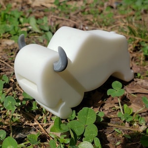 May include: A white, bull-shaped toilet paper holder with gray horns. The holder is resting on a bed of green grass and clover. The design is a whimsical addition to any bathroom.