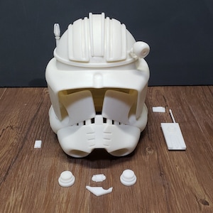 May include: A white, unassembled helmet with several separate parts. The helmet features a rounded top with vertical ridges and a visor. Additional components include two circular pieces, a rectangular part, and a small antenna.