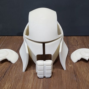 May include: A disassembled, off-white helmet with matching shoulder pieces and a chin guard. The helmet has a futuristic design with angular features and is made of a rigid material. The pieces are arranged on a wooden surface.