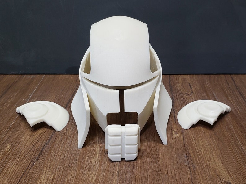 May include: A disassembled, off-white helmet with matching shoulder pieces and a chin guard. The helmet has a futuristic design with angular features and is made of a rigid material. The pieces are arranged on a wooden surface.