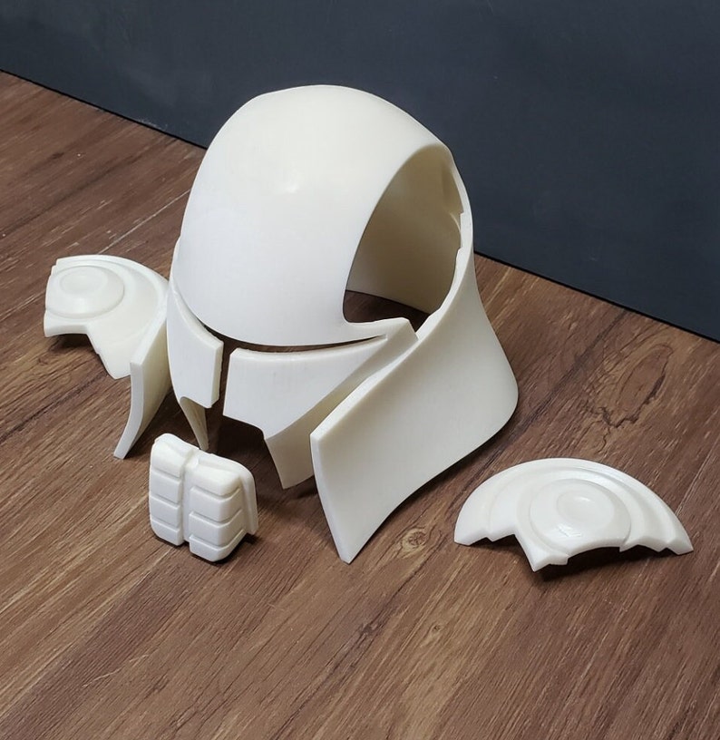 May include: Off-white, unassembled helmet parts. The components include a rounded top, side pieces, and smaller details. The pieces are arranged on a wooden surface, suggesting a DIY or cosplay project.
