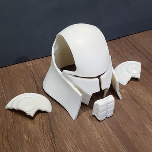May include: A disassembled white helmet, likely for cosplay or costume use, resting on a wooden surface. The helmet is composed of several separate pieces, including the main head covering, face guard, and side pieces.