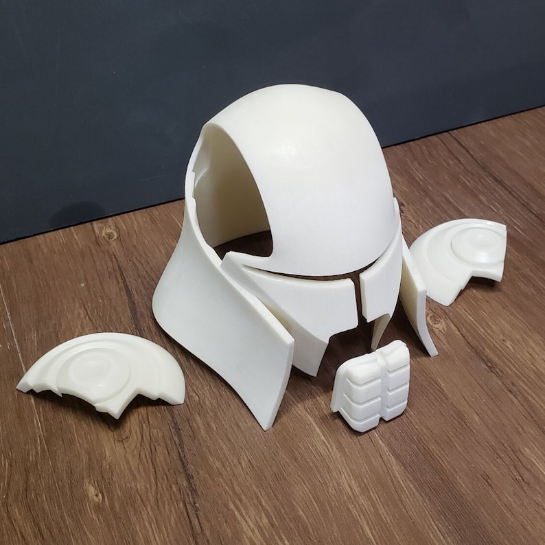 May include: A disassembled white helmet, likely for cosplay or costume use, resting on a wooden surface. The helmet is composed of several separate pieces, including the main head covering, face guard, and side pieces.