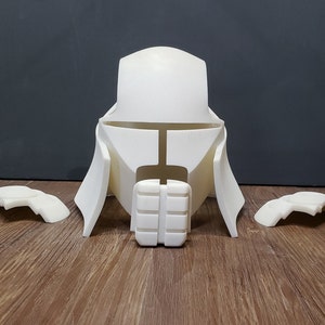 May include: A white, unassembled helmet and armor set, likely for cosplay or display. The set includes a central helmet piece with a distinctive shape, shoulder armor, and a chest piece. The pieces are made of a solid, opaque material.