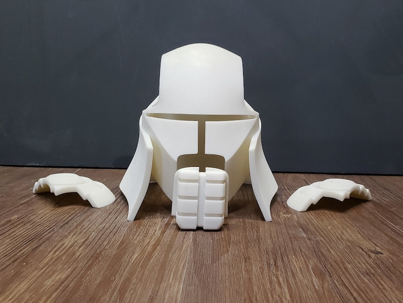 May include: A white, unassembled helmet and armor set, likely for cosplay or display. The set includes a central helmet piece with a distinctive shape, shoulder armor, and a chest piece. The pieces are made of a solid, opaque material.