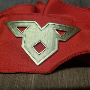 May include: A red fabric sash with a gold-colored metal emblem. The emblem features a central diamond shape with two triangles on either side. The sash is folded and lies on a wooden surface.