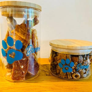Può includere: Due barattoli di vetro trasparente con coperchi in bambù, pieni di snack per cani. I barattoli hanno un motivo a impronta di zampa blu e il nome "Bonnie" in corsivo. Su una superficie di legno.