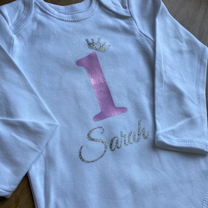 Peut inclure: Combinaison blanche à manches longues avec un numéro un rose et une couronne argentée pailletée. Le nom "Sarah" est écrit en argent pailleté sous le numéro.