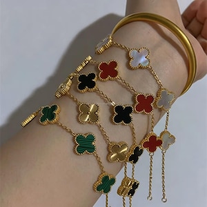 Peut inclure: Un bracelet jonc doré accompagné de plusieurs chaînes fines en or ornées de breloques en forme de trèfle. Les breloques sont incrustées de matériaux blancs, rouges, noirs et verts, formant un ensemble de bijoux superposés sur le poignet.