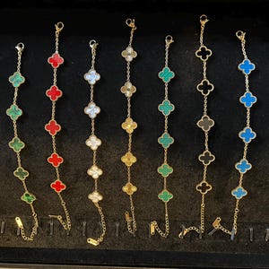 Peut inclure: Sept bracelets à chaîne dorée, chacun orné de cinq breloques en forme de trèfle de différentes couleurs, notamment vert, rouge, blanc, jaune, vert rayé, noir et bleu. Chaque bracelet comprend un fermoir mousqueton et une chaînette réglable.