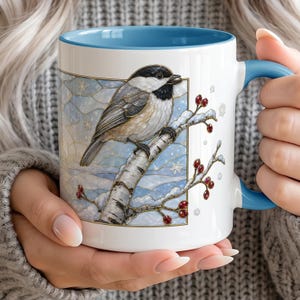 May include: A white ceramic mug with a blue interior and handle, featuring a graphic illustration of a black-capped chickadee perched on a snowy birch branch with red berries against a light blue winter background with snowflakes.