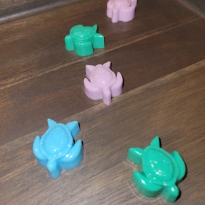 May include: Five small turtle-shaped figurines on a wooden surface. The turtles are in shades of green, blue, and purple.