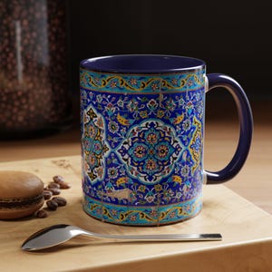Puede incluir: Una taza de cerámica con asa e interior azules, con un detallado y colorido estampado floral en tonos azules, turquesas, amarillos y blancos. La taza está sobre una superficie de madera con una cuchara y un pastel.