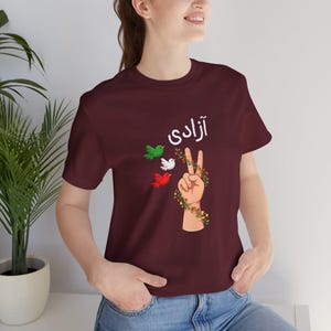 Unisex T-shirt featuring the Persian word “Azadi” meaning freedom, a hand making a peace sign wrapped in floral vines, and three doves in green, white, and red inspired by Iranian colors. Symbolic Iran freedom shirt representing peace, unity, and Woman Life Freedom movement.