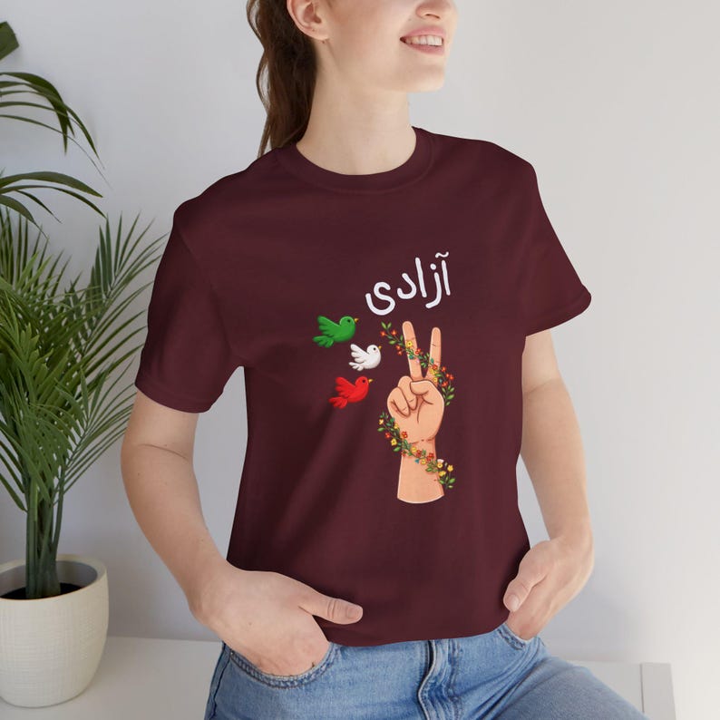 Unisex T-shirt featuring the Persian word “Azadi” meaning freedom, a hand making a peace sign wrapped in floral vines, and three doves in green, white, and red inspired by Iranian colors. Symbolic Iran freedom shirt representing peace, unity, and Woman Life Freedom movement.