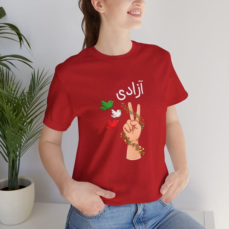Unisex T-shirt featuring the Persian word “Azadi” meaning freedom, a hand making a peace sign wrapped in floral vines, and three doves in green, white, and red inspired by Iranian colors. Symbolic Iran freedom shirt representing peace, unity, and Woman Life Freedom movement.