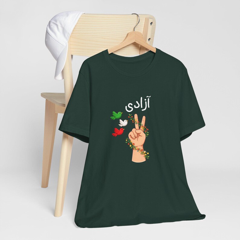 Unisex T-shirt featuring the Persian word “Azadi” meaning freedom, a hand making a peace sign wrapped in floral vines, and three doves in green, white, and red inspired by Iranian colors. Symbolic Iran freedom shirt representing peace, unity, and Woman Life Freedom movement.