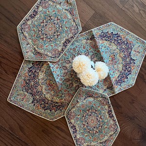 Può includere: Un set di quattro tovagliette decorative con un design a tappeto persiano nei toni del verde acqua, blu navy e ruggine. Le tovagliette hanno una forma geometrica unica e un bordo bianco. Un vaso di fiori bianchi è al centro.