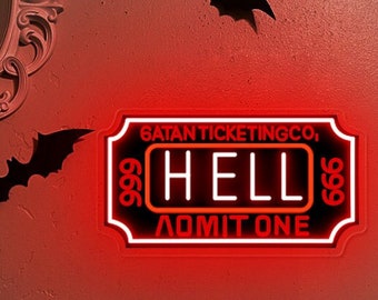 Op de afbeelding: Een rechthoekig neonbord met een rode en witte gloed in de vorm van een ticket. De tekst luidt SATAN TICKETING CO, HELL en ADMIT ONE, met het getal 666 aan beide kanten. Het bord is aan een muur bevestigd en geeft felrood licht af.