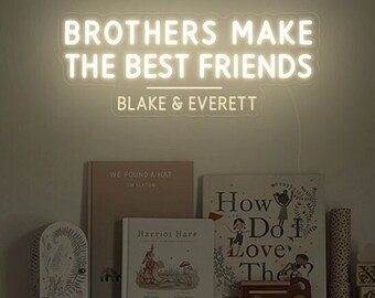 Op de afbeelding: Een warm wit neonbord aan de muur met de tekst BROTHERS MAKE THE BEST FRIENDS gevolgd door BLAKE & EVERETT. Onder het bord bevindt zich een houten plank met diverse kinderboeken, een kleine rode speelgoedvrachtwagen en een gevlochten mand.