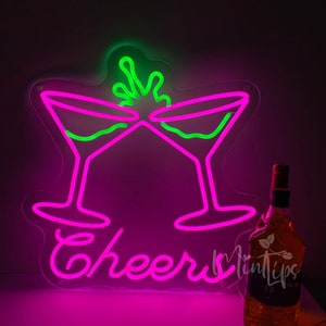 May include: A neon sign featuring two pink martini glasses clinking together with a green liquid splash above them and the word Cheers written in pink script below. A glass bottle of amber liquid stands to the right of the neon sign.