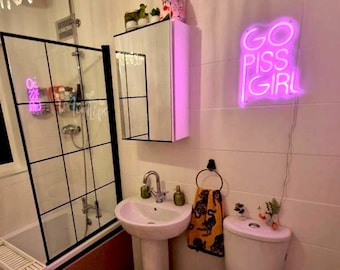 May include: A bathroom featuring a pink neon sign on the wall with the text GO PISS GIRL. The room includes a white pedestal sink, a toilet, a bathtub with a black-framed glass screen, a crystal chandelier, and a rug with moon phase patterns.