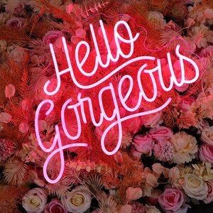 May include: A bright pink neon sign displaying the text Hello Gorgeous in a cursive script font. The sign is mounted against a dense background of artificial flowers in shades of cream, light pink, and dusty orange, including roses and ferns.