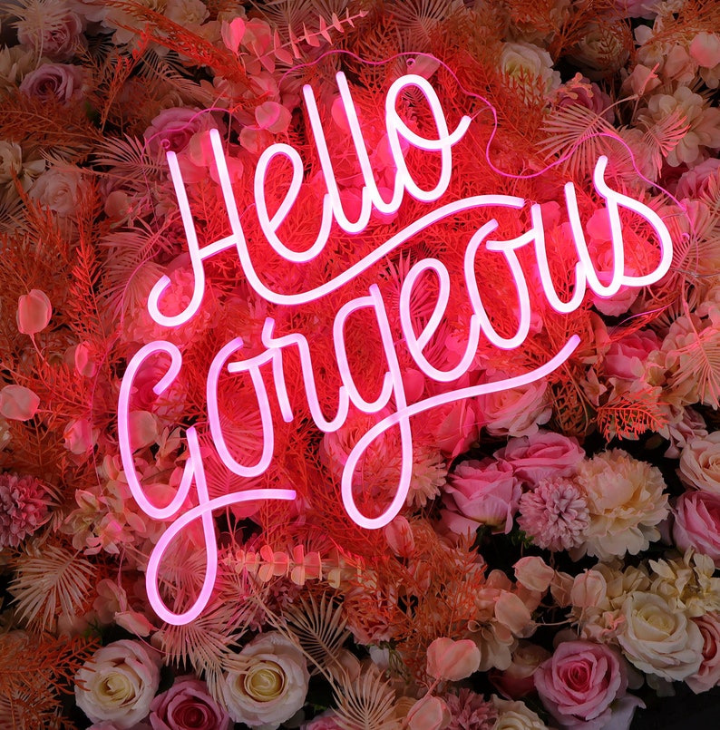 May include: A bright pink neon sign displaying the text Hello Gorgeous in a cursive script font. The sign is mounted against a dense background of artificial flowers in shades of cream, light pink, and dusty orange, including roses and ferns.