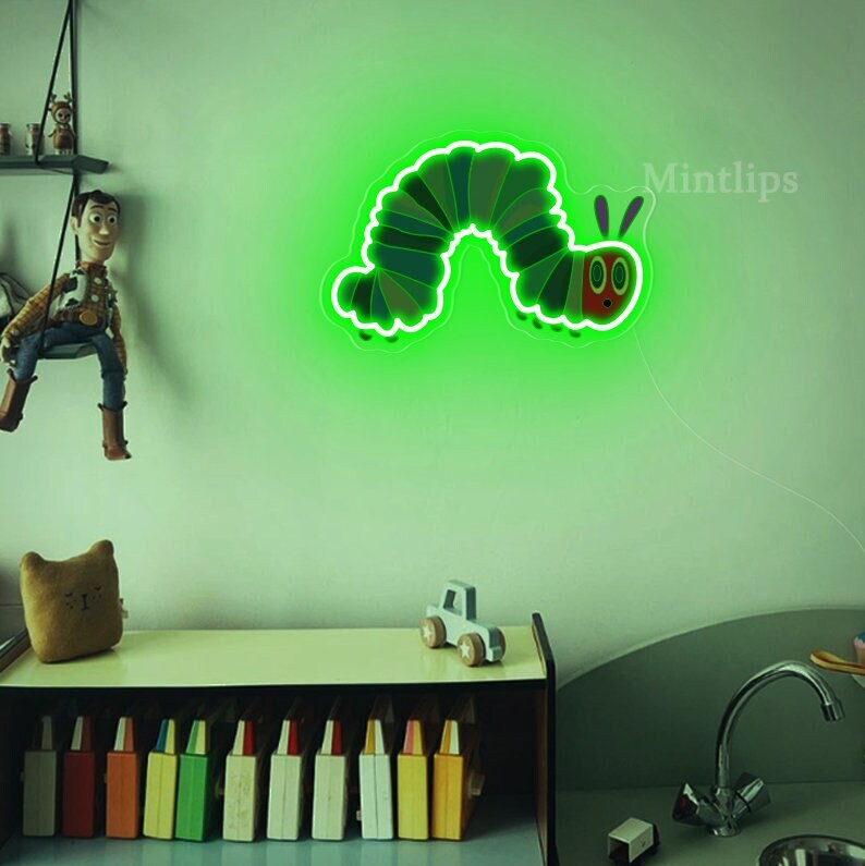 May include: Green neon light shaped like a caterpillar with a red face and two antennae. The light is mounted on a white wall in a room with a bookshelf and a toy car.