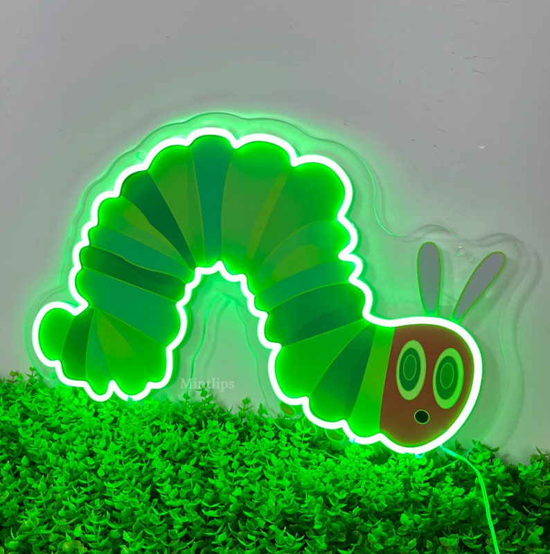 May include: A green neon sign shaped like a caterpillar with a red face and two large eyes. The sign is lit up and is sitting on a bed of green plants.