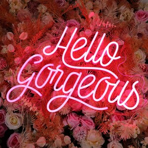 May include: A bright pink neon sign displaying the text Hello Gorgeous in a cursive script font. The sign is mounted against a dense backdrop of artificial flowers and foliage in shades of dusty rose, cream, peach, and muted orange tones.