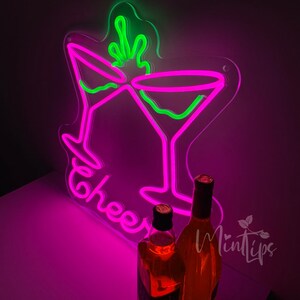 May include: A neon sign featuring two martini glasses in bright pink and green, with the word Cheers written in pink script below the glasses. Two glass bottles containing amber liquid are positioned in front of the glowing neon light display.