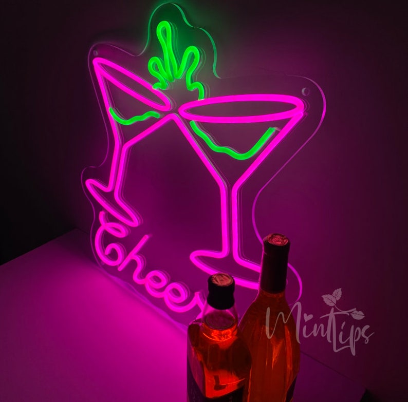 May include: A neon sign featuring two martini glasses in bright pink and green, with the word Cheers written in pink script below the glasses. Two glass bottles containing amber liquid are positioned in front of the glowing neon light display.