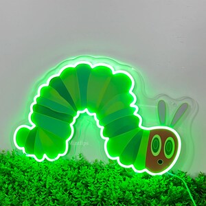 May include: Green neon sign of a caterpillar with a smiling face. The caterpillar is curled into a circle shape. The sign is on a white background with green plants in the foreground.