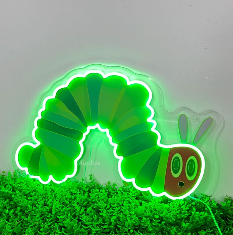 May include: Green neon sign of a caterpillar with a smiling face. The caterpillar is curled into a circle shape. The sign is on a white background with green plants in the foreground.