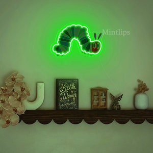 May include: Green neon light shaped like a caterpillar with a red face and antennae. The light is mounted on a wall above a shelf with a book, a small cabinet, and a vase.