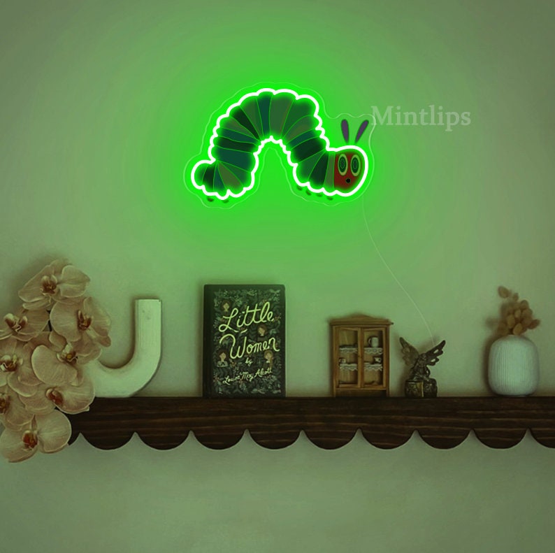 May include: Green neon light shaped like a caterpillar with a red face and antennae. The light is mounted on a wall above a shelf with a book, a small cabinet, and a vase.