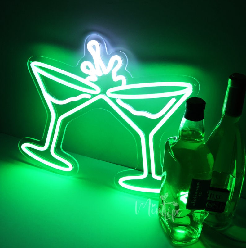May include: A bright green neon sign depicting two martini glasses clinking together with a splash effect above them. The sign emits a vibrant green glow, illuminating two glass bottles positioned in the foreground to the right of the neon light.