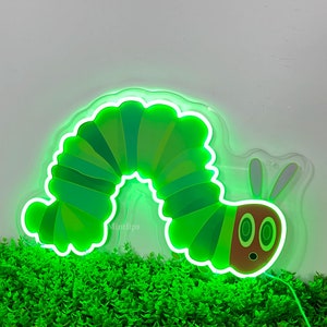 May include: A green neon sign shaped like a caterpillar with a brown head and two large eyes. The sign is lit up and is sitting on a bed of green plants.