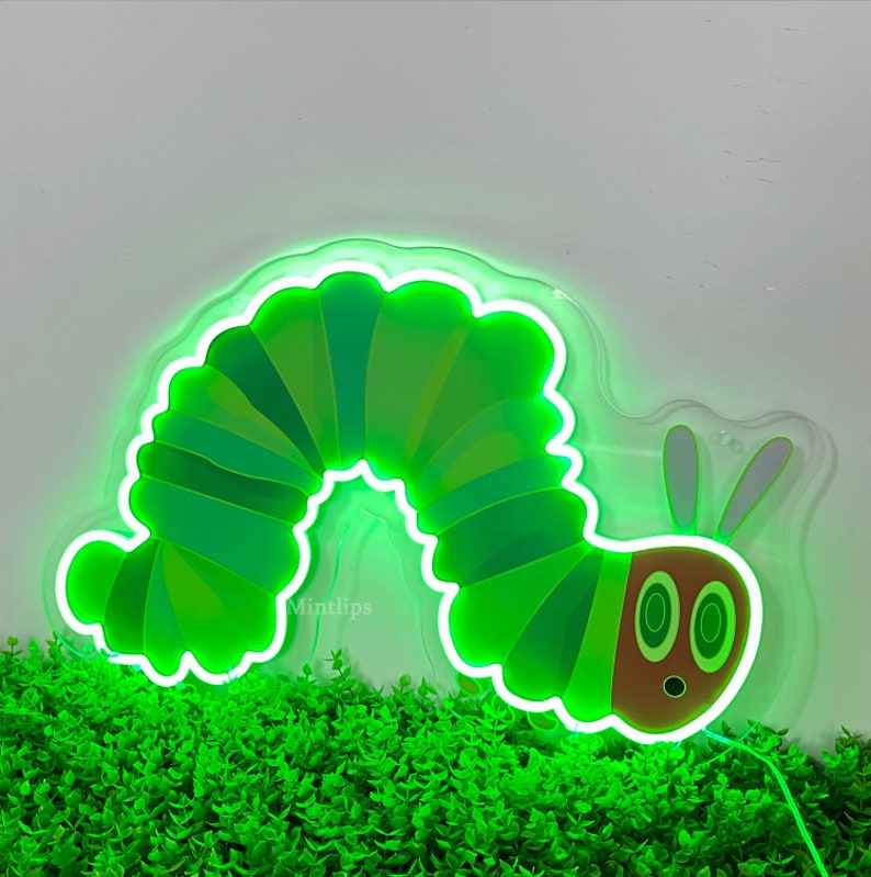 May include: A green neon sign shaped like a caterpillar with a brown head and two large eyes. The sign is lit up and is sitting on a bed of green plants.