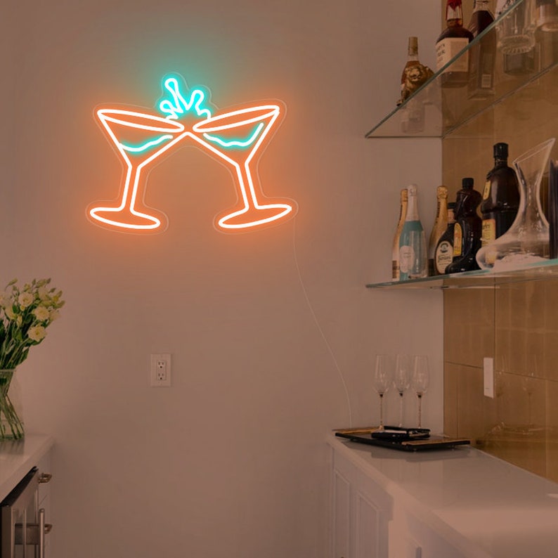 May include: A neon sign mounted on a wall depicting two martini glasses clinking together in a toast. The outline of the glasses is bright orange, while the liquid inside and the splash effect at the top are illuminated in a vibrant teal color.