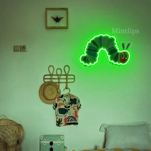 May include: A green neon light shaped like a caterpillar with a red face and two antennae. The light is mounted on a white wall with a wooden picture frame, a hat rack, and a hat hanging on it. The text "Mintlips" is visible in the top right corner of the image.