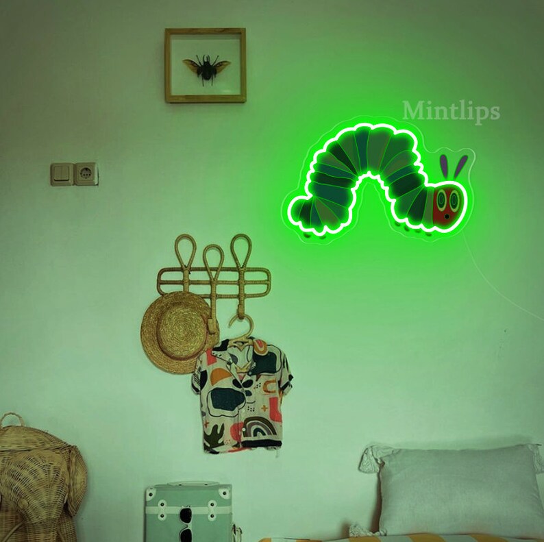 May include: A green neon light shaped like a caterpillar with a red face and two antennae. The light is mounted on a white wall with a wooden picture frame, a hat rack, and a hat hanging on it. The text "Mintlips" is visible in the top right corner of the image.